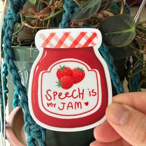 May include: A sticker in the shape of a jam jar with a red and white checkered lid. The jar is filled with red jam and has a label with three strawberries and the text "Speech is my JAM" with a heart.