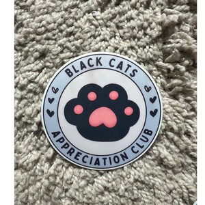 May include: A round sticker with a light blue border and the text "BLACK CATS APPRECIATION CLUB". The sticker features a black cat paw print with pink pads and small hearts.