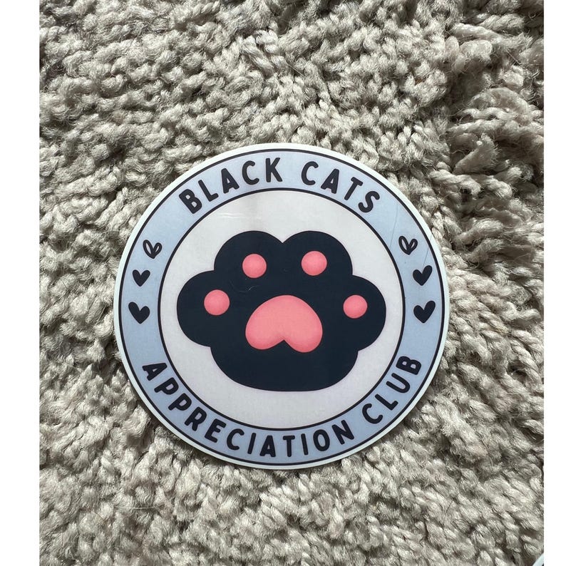 May include: A round sticker with a light blue border and the text "BLACK CATS APPRECIATION CLUB". The sticker features a black cat paw print with pink pads and small hearts.