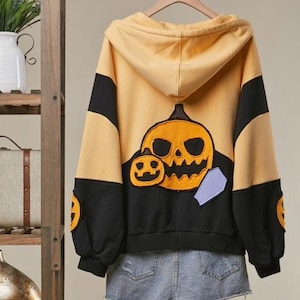 May include: A yellow and black hooded sweatshirt featuring a large orange jack-o'-lantern patch on the back, smaller pumpkin patches on the sleeves, and a light purple coffin shape. The hoodie is paired with light blue denim shorts.
