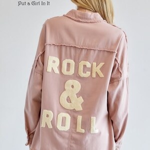 May include: A dusty rose-colored jacket with the words "ROCK & ROLL" appliquéd on the back in a cream-colored, block-letter font. The jacket has a collared neckline and long sleeves. The text "Put a Girl In It" is printed on the upper left.