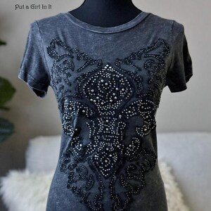 May include: Dark gray t-shirt with a decorative black beaded design on the front. The design is an ornate pattern with small, sparkling embellishments. The text "Put a Girl In It" is visible at the top.