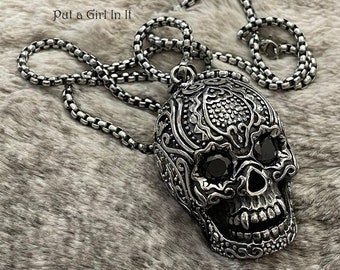 May include: A silver chain necklace with a detailed skull pendant. The skull has black eyes and intricate designs on the face. The pendant is about 2 inches tall.