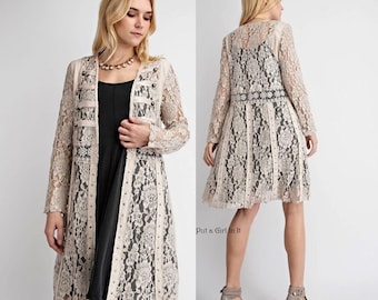 May include: A long, beige lace cardigan with a black lace trim and a black and white beaded embellishment. The cardigan has a fitted bodice and a flowing skirt. The model is wearing a black dress underneath the cardigan.