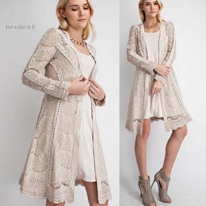 May include: A beige lace duster coat with long sleeves and a scalloped hemline. The coat is worn over a cream-colored dress. The text "Put a Girl In It" is in the upper left corner. The coat features decorative studs.