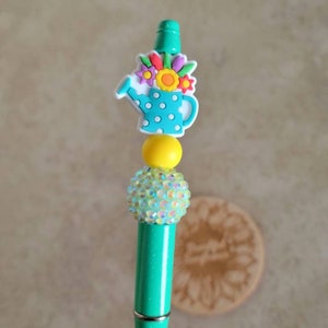 May include: A turquoise pen with a green top and a white watering can charm with colorful flowers. The watering can has white polka dots. The pen has a yellow bead and a sparkly clear bead.