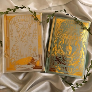 Op de afbeelding: Twee hardcover boeken met goudfolie details. Het linkerboek is wit met een illustratie van een figuur en een leeuw getiteld Circe. Het rechterboek is lichtblauw met een lier en een gezicht getiteld The Song of Achilles. Beide hebben gouden hoeken.