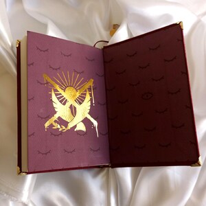 Puede incluir: Un diario encuadernado en cuero burdeos con detalles dorados y un dise&ntilde;o en relieve dorado en la portada. El dise&ntilde;o presenta dos palomas con ramas de olivo y un sol con rayos. La contraportada tiene un patr&oacute;n repetitivo de pesta&ntilde;as doradas.