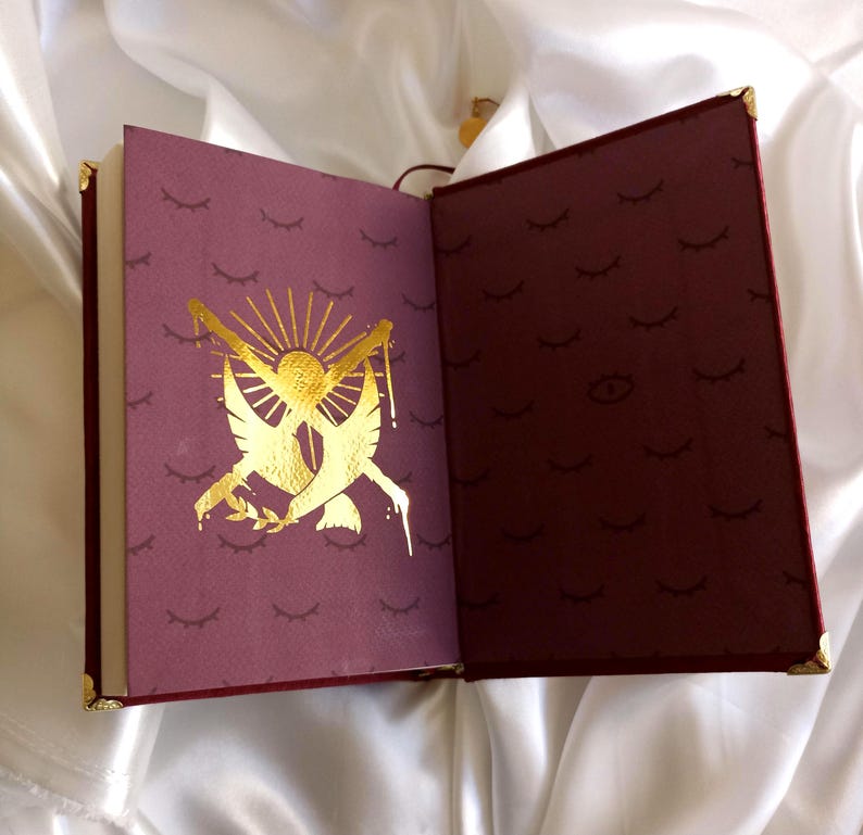 Puede incluir: Un diario encuadernado en cuero burdeos con detalles dorados y un dise&ntilde;o en relieve dorado en la portada. El dise&ntilde;o presenta dos palomas con ramas de olivo y un sol con rayos. La contraportada tiene un patr&oacute;n repetitivo de pesta&ntilde;as doradas.