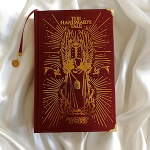 Puede incluir: Un libro de tapa dura rojo con letras e ilustraciones doradas. El t&iacute;tulo es "El cuento de la criada" de Margaret Atwood.