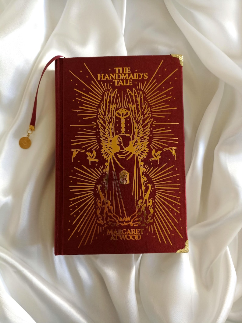 Puede incluir: Un libro de tapa dura rojo con letras e ilustraciones doradas. El t&iacute;tulo es "El cuento de la criada" de Margaret Atwood.