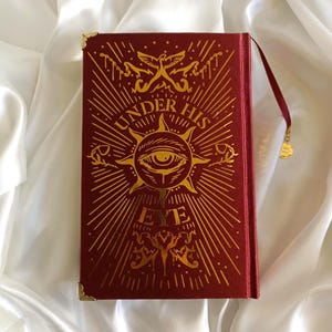 Puede incluir: Un libro de tapa dura rojo con letras doradas y un dise&ntilde;o dorado en la portada. El t&iacute;tulo del libro es "Under His Eye".