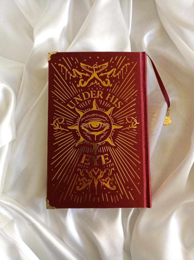 Puede incluir: Un libro de tapa dura rojo con letras doradas y un dise&ntilde;o dorado en la portada. El t&iacute;tulo del libro es "Under His Eye".