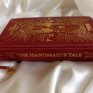 Puede incluir: Un libro de tapa dura rojo con letras doradas y un dise&ntilde;o dorado en la portada. El t&iacute;tulo del libro es "The Handmaid's Tale".