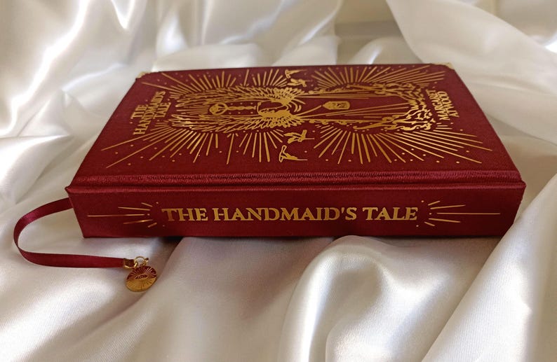 Puede incluir: Un libro de tapa dura rojo con letras doradas y un dise&ntilde;o dorado en la portada. El t&iacute;tulo del libro es "The Handmaid's Tale".