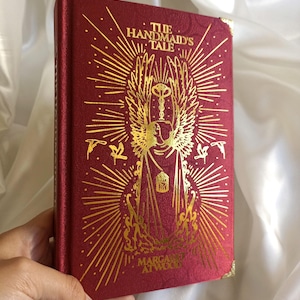 Puede incluir: Un libro de tapa dura rojo con letras doradas y una ilustraci&oacute;n dorada de una figura alada sosteniendo una llave. El t&iacute;tulo del libro es "El cuento de la criada" de Margaret Atwood.