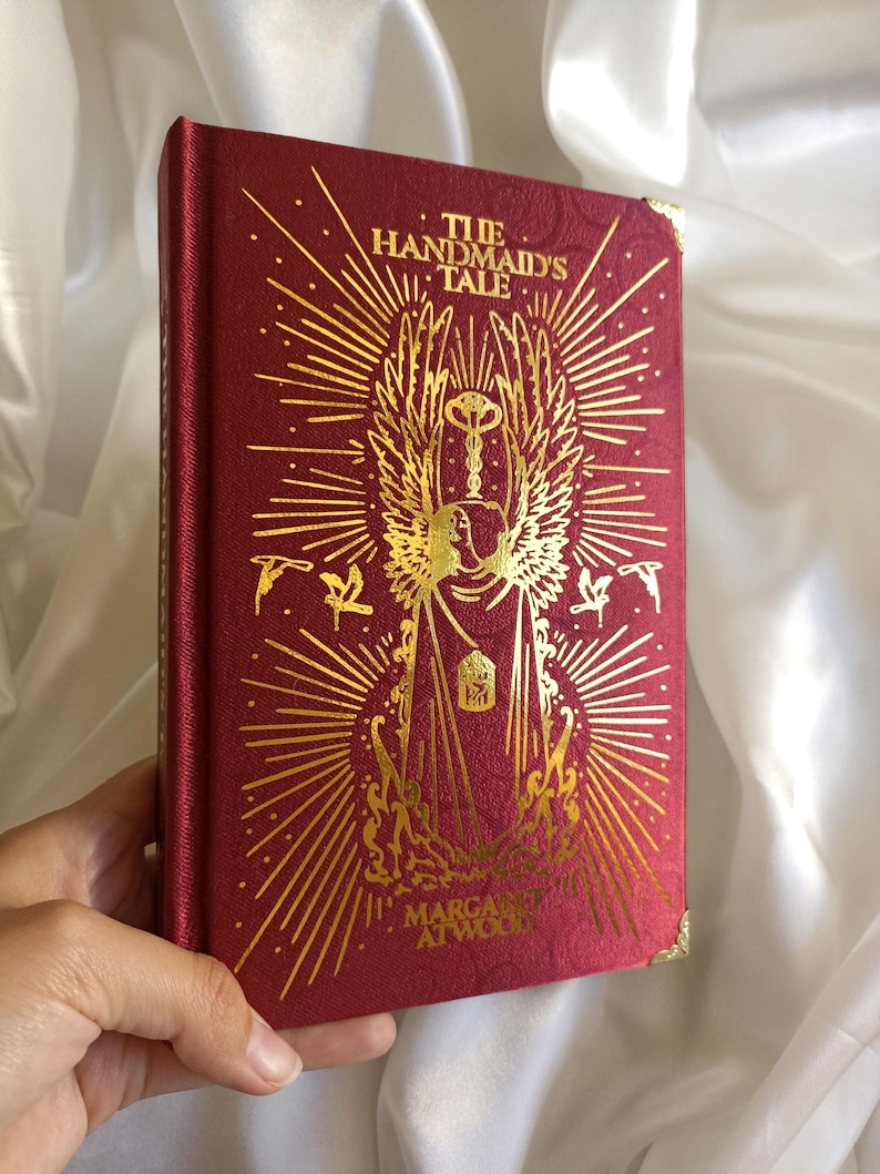 Puede incluir: Un libro de tapa dura rojo con letras doradas y una ilustraci&oacute;n dorada de una figura alada sosteniendo una llave. El t&iacute;tulo del libro es "El cuento de la criada" de Margaret Atwood.