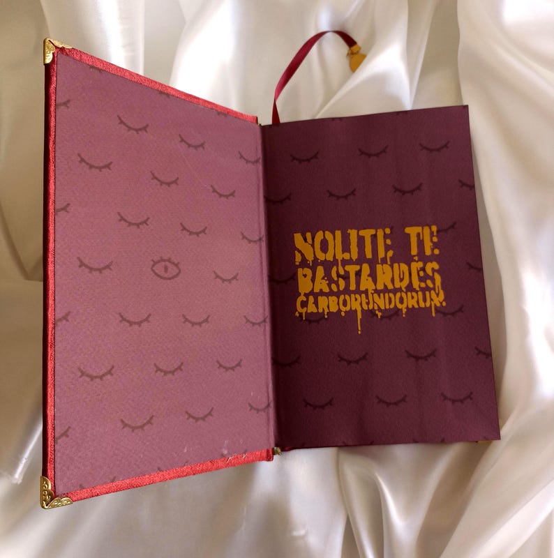 Puede incluir: Un libro color burdeos con un cierre dorado y un marcap&aacute;ginas de cinta roja. La portada tiene un patr&oacute;n de ojos y pesta&ntilde;as. La contraportada tiene el texto "NOLITE TE BASTARDES CARBORUNDORUM" en letras doradas.