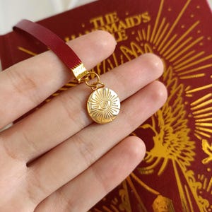 Puede incluir: Un marcap&aacute;ginas de sat&eacute;n rojo con un dije dorado que presenta un ojo estilizado con rayos de sol. El dije est&aacute; unido al marcap&aacute;ginas con un anillo dorado.