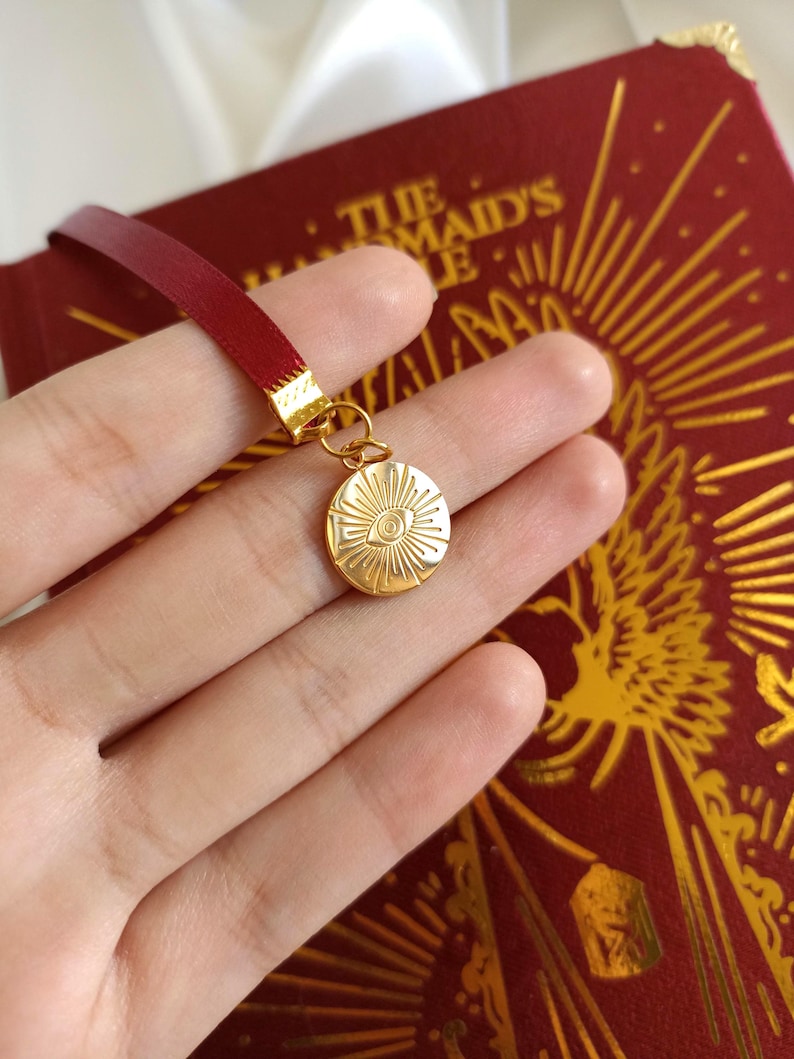 Puede incluir: Un marcap&aacute;ginas de sat&eacute;n rojo con un dije dorado que presenta un ojo estilizado con rayos de sol. El dije est&aacute; unido al marcap&aacute;ginas con un anillo dorado.