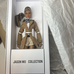 May include: A fashion doll from the Jason Wu Collection, presented in a white box. The doll wears a tan trench coat, white shirt, shorts, and a black and white polka dot bow tie. The doll has brown hair and is in a box with the text "JASON WU COLLECTION".
