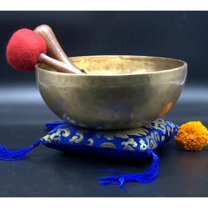 May include: A brass singing bowl with two wooden mallets, one with a red felt head, rests on a blue cushion with gold embroidery and tassels. A small orange flower sits nearby. The bowl is a meditation tool.