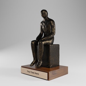 Puede incluir: Una escultura de tono bronce de una figura humana sentada con una superficie texturizada, colocada sobre un bloque cuadrado. La escultura descansa sobre una base rectangular de madera con una placa clara con el texto grabado Your Text Here.