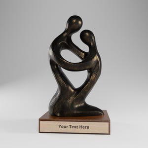 Peut inclure: Une sculpture abstraite de couleur bronze représentant deux silhouettes enlacées, fixée sur un socle rectangulaire en bois. Le socle comporte une plaque de couleur claire avec le texte Your Text Here centré sur la face avant du support.
