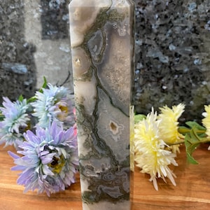 May include: A green and grey agate crystal point, also known as a wand or obelisk, with a pointed tip and a flat base. The crystal has a natural, organic pattern of green and grey swirls and veins.