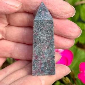May include: A rectangular, grey and red crystal obelisk with a pointed top. The stone has a speckled appearance with red and grey tones. The obelisk is held in a person's hands, with a blurred background of green foliage and pink flowers.