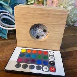 May include: A white remote control with 16 colour buttons, a white 'W' button, a red 'ON' button, a black 'OFF' button, and mode buttons labelled 'Flash', 'Strobe', 'Fade', and 'Smooth'. The remote is next to a wooden base with a round cutout for a light.