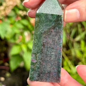 May include: A green and dark red crystal obelisk with a pointed top. The crystal has a textured surface and is held in a person's hand. The background is blurred with green foliage.