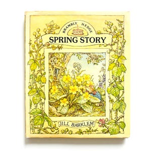 May include: A hardcover book titled Brambly Hedge Spring Story by Jill Barklem. The cover features a yellow background with a central illustration of a mouse among yellow primrose flowers, framed by detailed green vines and thorny stems.