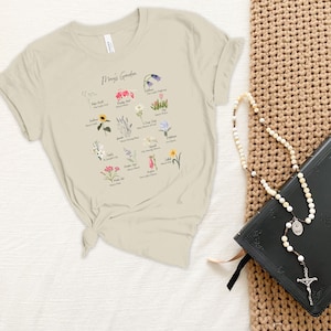 Op de afbeelding: Een beige T-shirt met een afbeelding van een bloemenkaart met de titel "Mary's Garden". De kaart toont een verscheidenheid aan bloemen met hun namen onder elke afbeelding. Het T-shirt is aan de voorkant geknoopt en ligt op een wit oppervlak. Een zwart lederen gebonden boek en een witte rozenkrans liggen op een bruin geweven oppervlak.