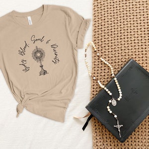 May include: A beige t-shirt with the text "God's Blood, Soul & Divinity" and a graphic of a monstrance. A black leather-bound book and a white rosary are on a brown woven surface.