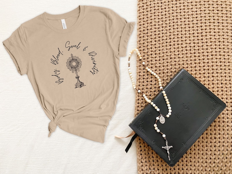 May include: A beige t-shirt with the text "God's Blood, Soul & Divinity" and a graphic of a monstrance. A black leather-bound book and a white rosary are on a brown woven surface.