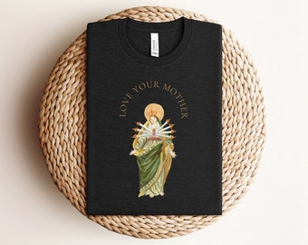 以下が含まれることがあります： 金のグラフィックが施された黒いTシャツ。グラフィックには、王冠をかぶった女性が描かれており、彼女の心臓には7本の剣が突き刺さっています。グラフィックの周りを「LOVE YOUR MOTHER」という文字が囲んでいます。