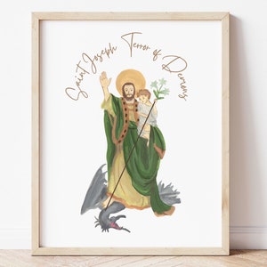 Può includere: Stampa ad acquerello incorniciata di San Giuseppe, con aureola, che tiene in braccio un bambino e un bastone, in piedi su un drago grigio scuro. Il testo "Saint Joseph Terror of Demons" è arcuato sopra l'immagine in oro. La cornice è in legno chiaro.
