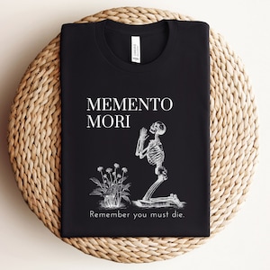 Puede incluir: Camiseta negra con la frase "Memento Mori" y una ilustraci&oacute;n de un esqueleto arrodillado rezando, junto a una planta. El texto "Remember you must die" est&aacute; debajo de la ilustraci&oacute;n.