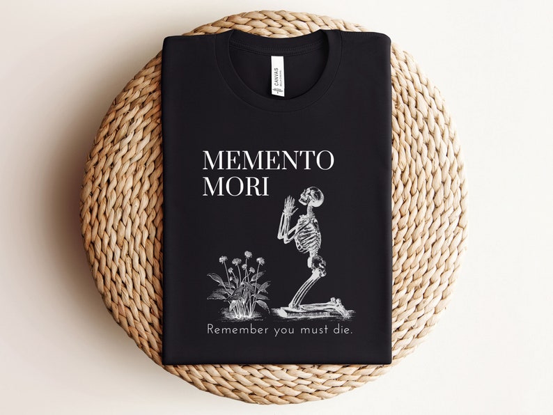 Puede incluir: Camiseta negra con la frase "Memento Mori" y una ilustraci&oacute;n de un esqueleto arrodillado rezando, junto a una planta. El texto "Remember you must die" est&aacute; debajo de la ilustraci&oacute;n.