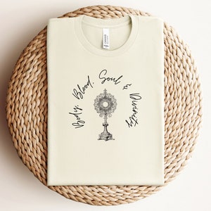 May include: A cream-coloured t-shirt with a black graphic of a monstrance and the text "Body, Blood, Soul & Divinity".