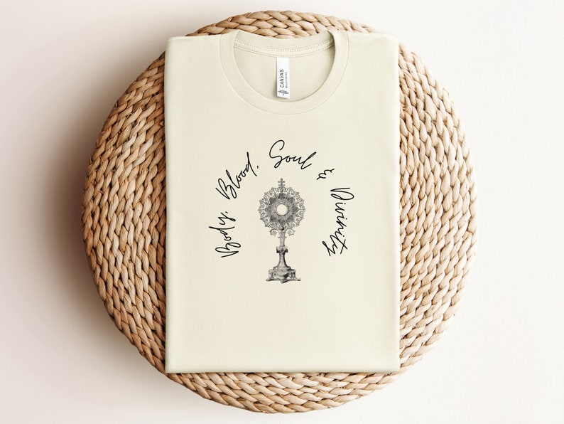 May include: A cream-coloured t-shirt with a black graphic of a monstrance and the text "Body, Blood, Soul & Divinity".