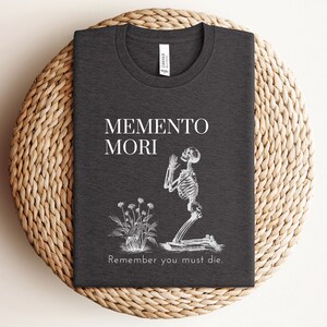 Puede incluir: Camiseta gris oscuro con las palabras "MEMENTO MORI" y "Remember you must die." Se representa un esqueleto arrodillado y un peque&ntilde;o arreglo floral. La camiseta est&aacute; doblada sobre una superficie circular tejida.