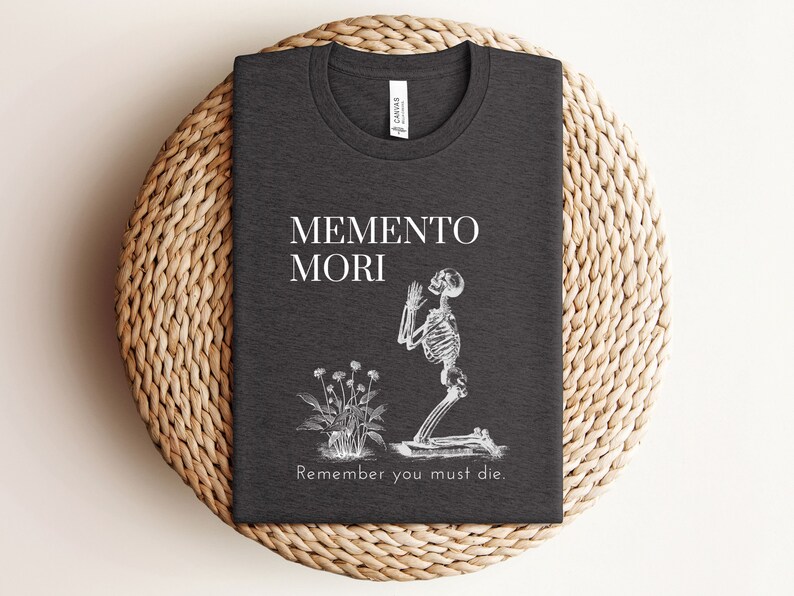 Puede incluir: Camiseta gris oscuro con las palabras "MEMENTO MORI" y "Remember you must die." Se representa un esqueleto arrodillado y un peque&ntilde;o arreglo floral. La camiseta est&aacute; doblada sobre una superficie circular tejida.