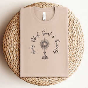 May include: A light brown t-shirt with a black graphic of a monstrance and the text "Body, Blood, Soul & Divinity".