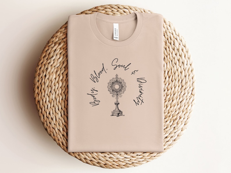May include: A light brown t-shirt with a black graphic of a monstrance and the text "Body, Blood, Soul & Divinity".
