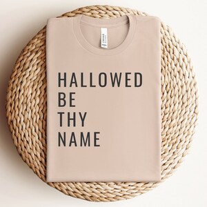 May include: A light brown t-shirt with the text "HALLOWED BE THY NAME" printed in black.