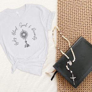 May include: White t-shirt featuring a black and white image of a monstrance with the text "Body, Blood, Soul & Divinity". A black leather-bound book and a white rosary with a silver cross are on a brown woven surface.