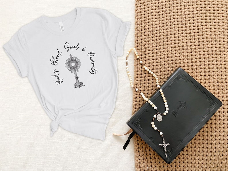 May include: White t-shirt featuring a black and white image of a monstrance with the text "Body, Blood, Soul & Divinity". A black leather-bound book and a white rosary with a silver cross are on a brown woven surface.