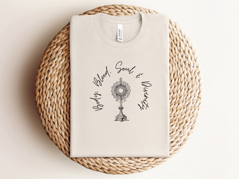 May include: A beige t-shirt with the text "Body, Blood, Soul & Divinity" printed in black cursive script. A black and white image of a monstrance is centred on the shirt.